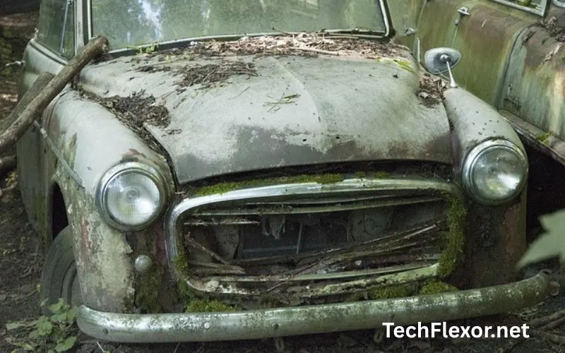 Old scrap car parked in backyard lawn with flat tires, covered in dust and fallen tree leaves