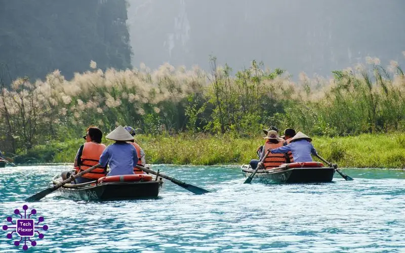 People rowing small boats across a calm river surrounded by green vegetation and misty hills